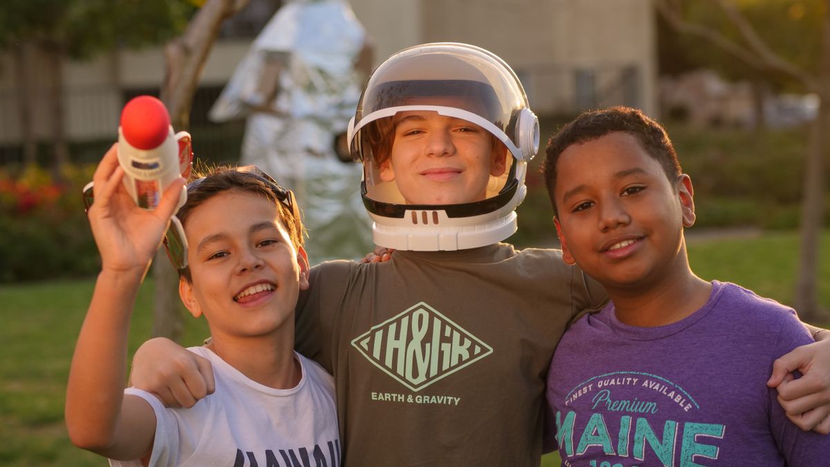 Three happy children playing outdoors with a space helmet and toy rocket on a sunny day.