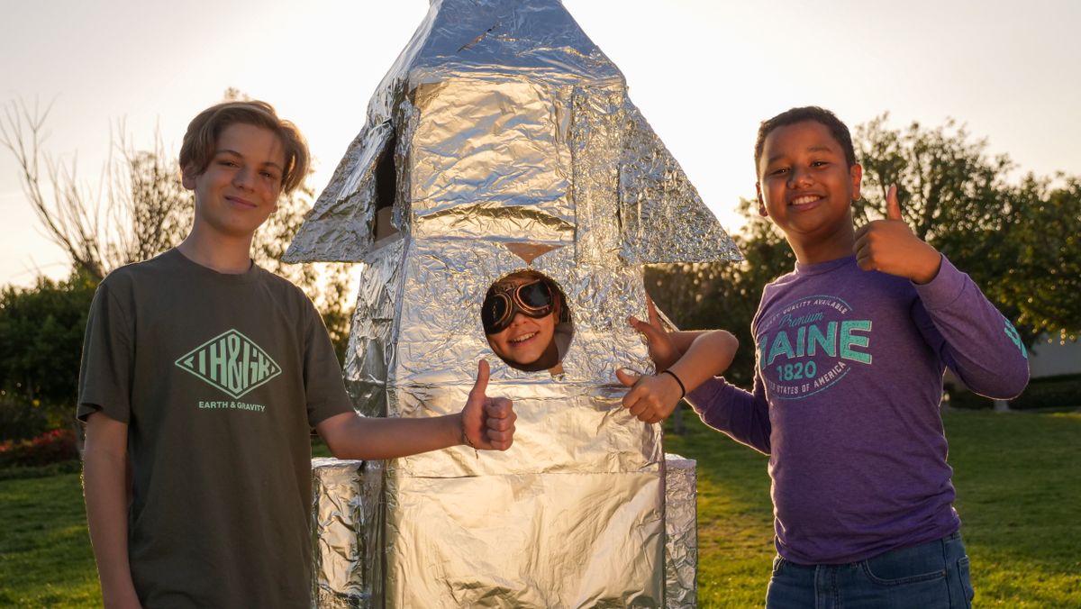 Three kids enjoy outdoor play with a handmade foil rocket on a sunny day.