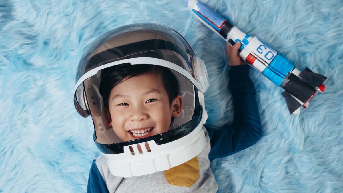 Happy child wearing an astronaut helmet lying on a blue rug, holding a toy rocket.