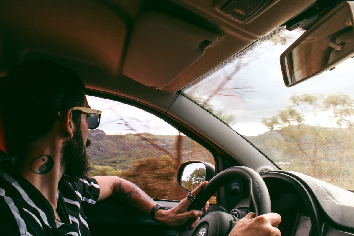 Bearded man with tattoo driving a car through scenic Diamantina, Brazil, wearing sunglasses.