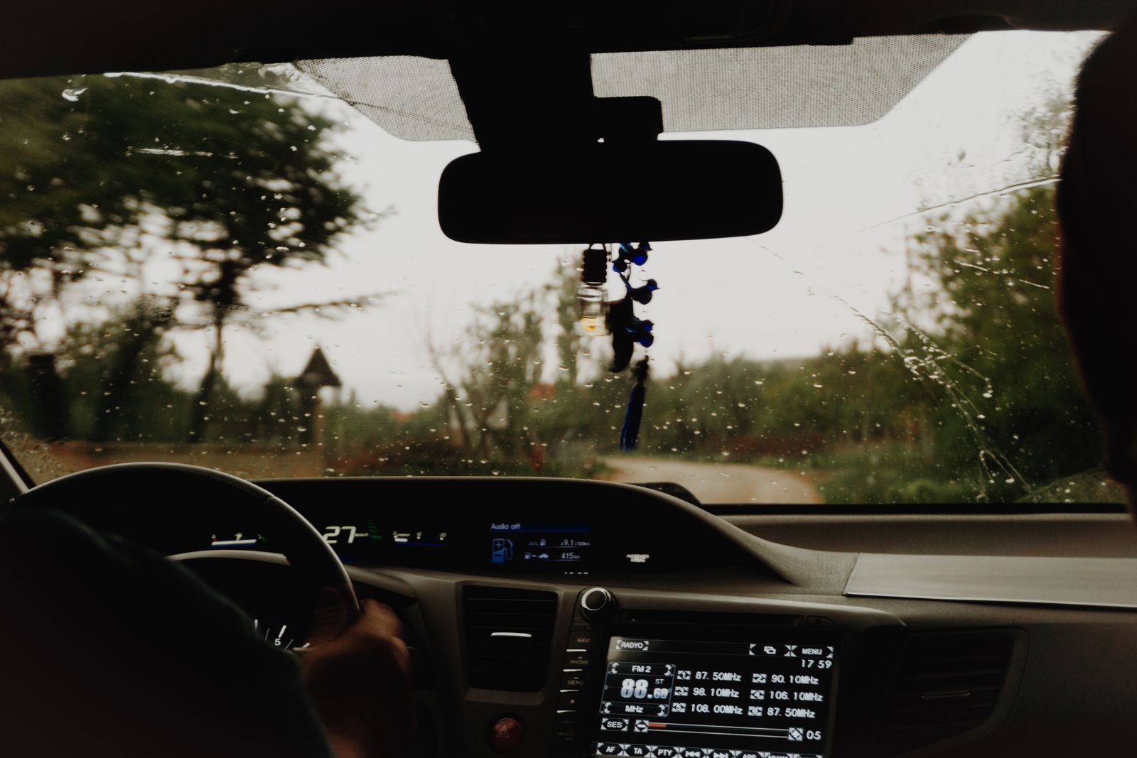 View from a car interior during a rainy drive on a rural road. Steering wheel and dashboard visible.