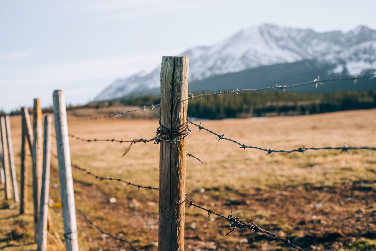 A rustic barbed wire fence in a sunny rural landscape with mountains in the background.