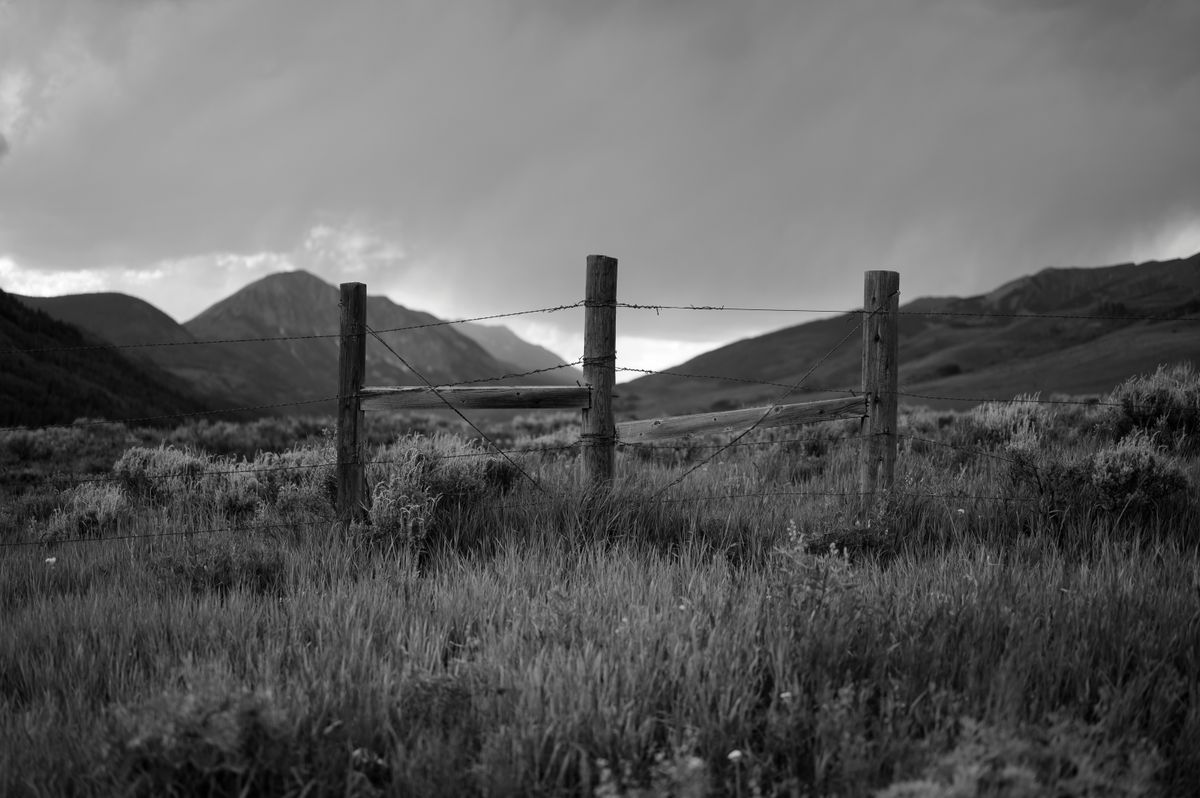 Monochrome landscape featuring a wooden barbed wire fence and scenic mountains, evoking rural tranquility.