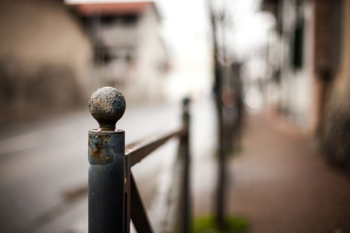 Close-up of a rusty metal fence post with a blurred urban street background depicting decay and urban life.