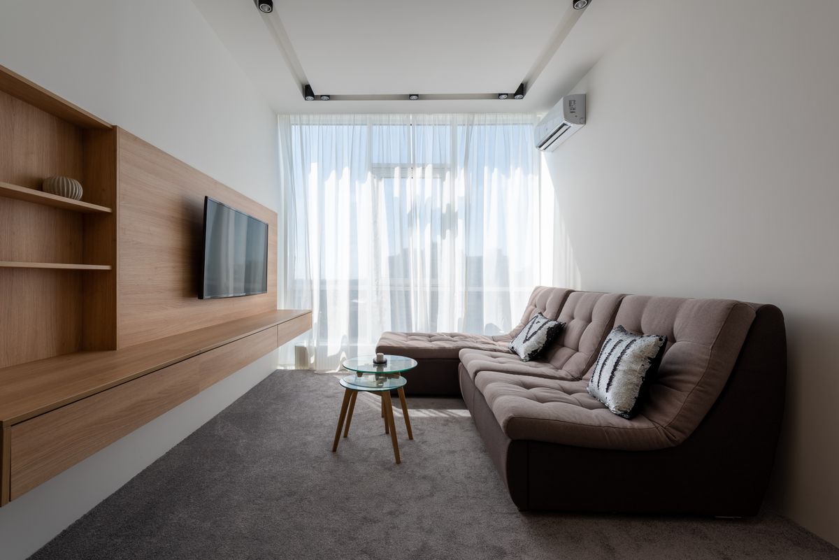 Comfortable couch with cushions near side tables against wall with wooden cupboard and modern TV in stylish living room with window