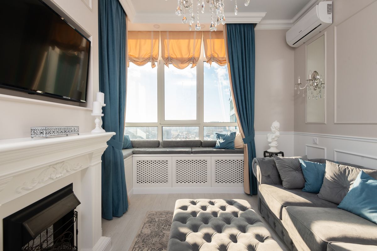 Comfortable sofa with cushions and pouf placed against wall with TV and fireplace in spacious stylish room with window and curtains