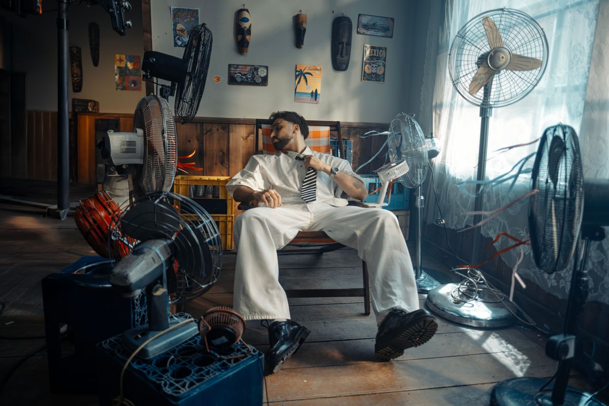 A man in white attire sits surrounded by various fans, cooling off in a warmly lit room.