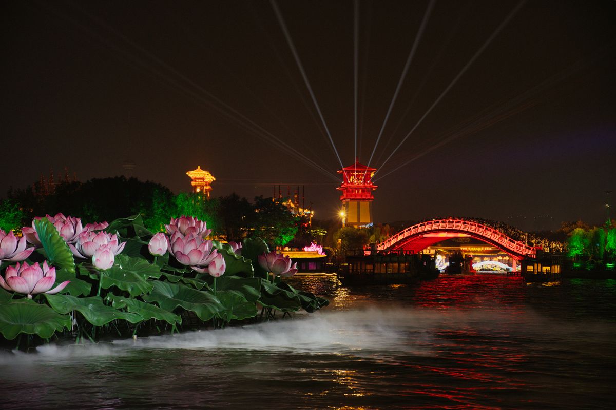 A spectacular night scene of a bridge with colorful lights and lotus flowers on a water body in Kaifeng, China.