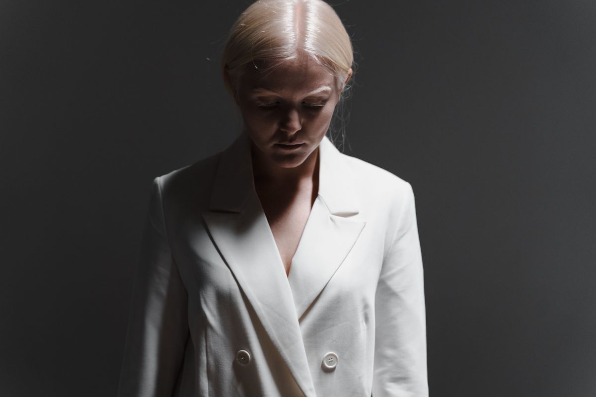 Studio portrait of a woman with blonde hair looking down, wearing a white coat.
