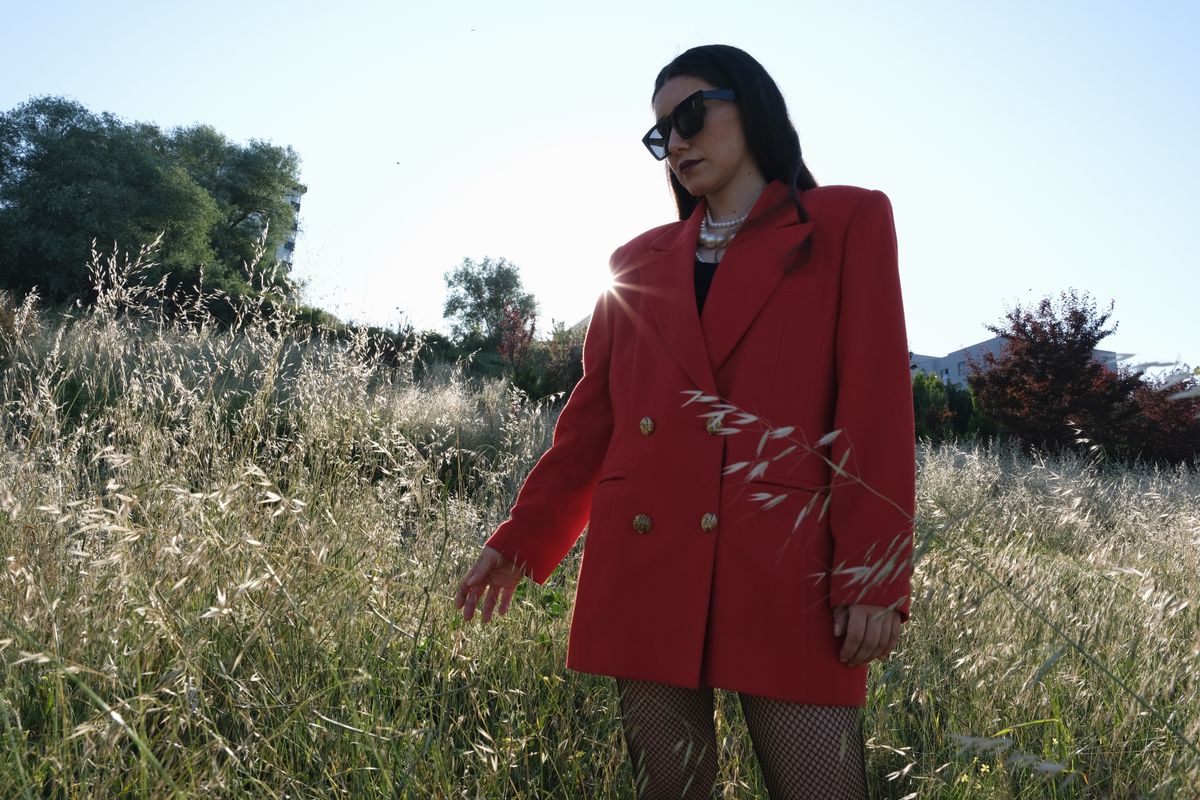 Fashionable woman wearing a red coat stands in a sunlit meadow, exuding elegance.