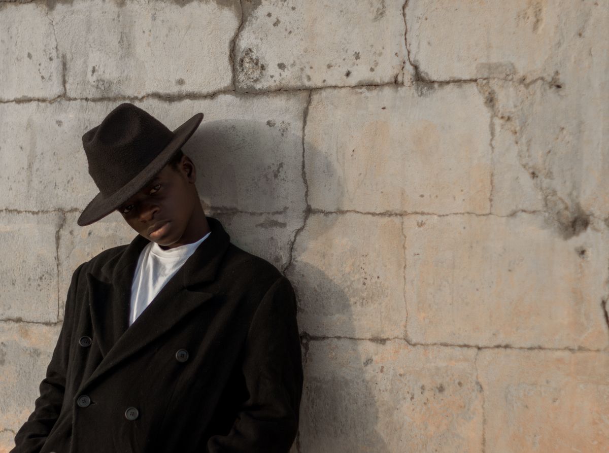Young man in a black coat and hat leans against a concrete wall, exuding style and attitude.