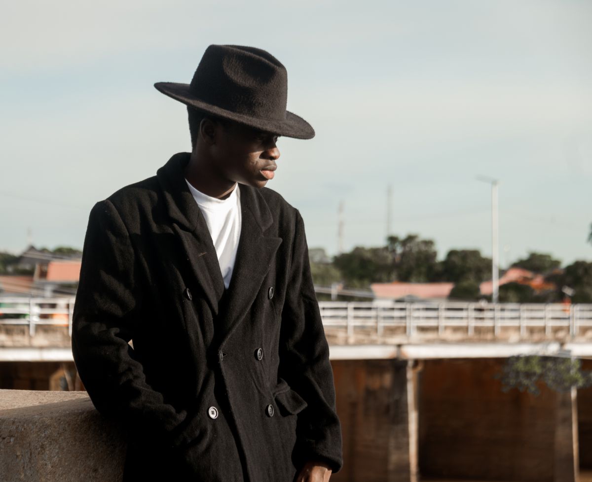 A fashionable man wearing a black coat and hat leans on a ledge outdoors against a bridge and city background.