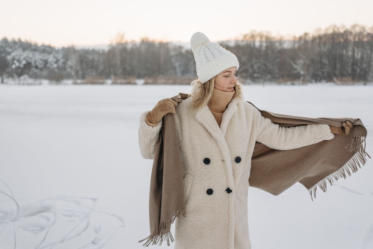 A woman in a warm winter coat stands by a snow-covered lake, adjusting her scarf.