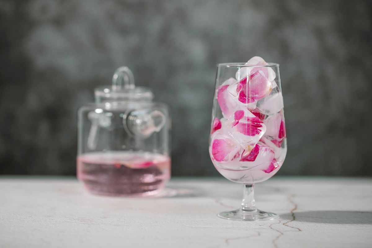 Transparent glass full of ice cubes and petals of pink rose placed on table near jar of refreshing drink