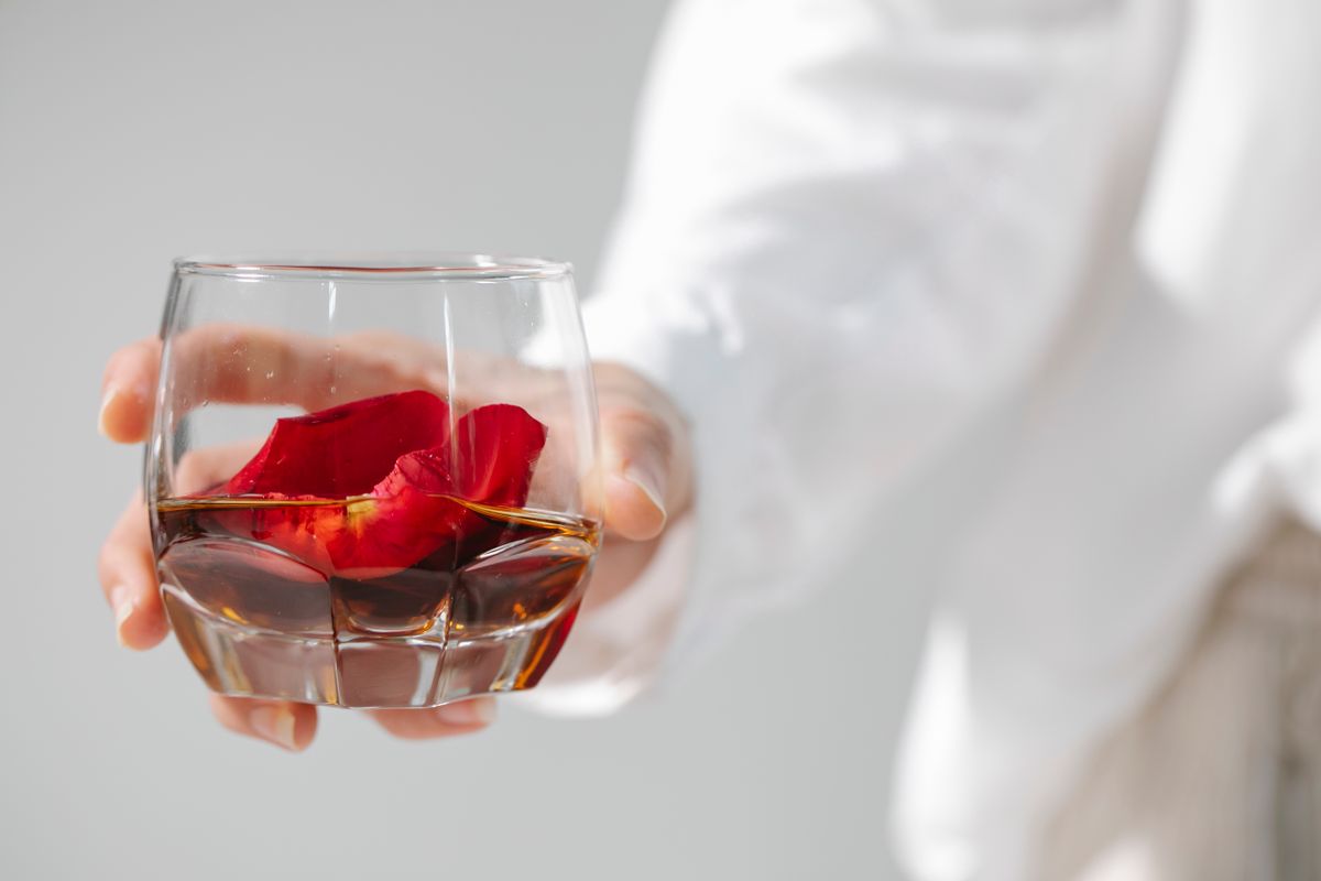 Crop faceless person in white shirt with transparent glass of alcoholic beverage with red rose petals