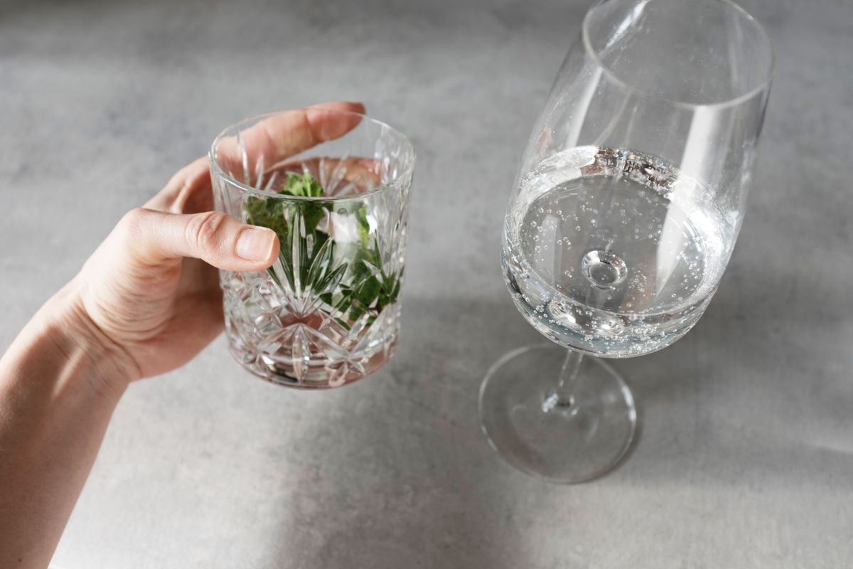 Unrecognizable bartender making healthy refreshing drink by adding mineral water from wineglass to faceted glass with herbs against gray board