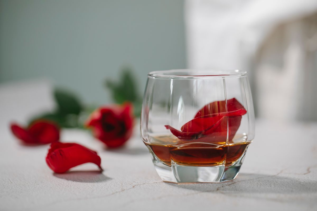 Crystal glass of alcoholic beverage with red petals of rose on table near rose flower