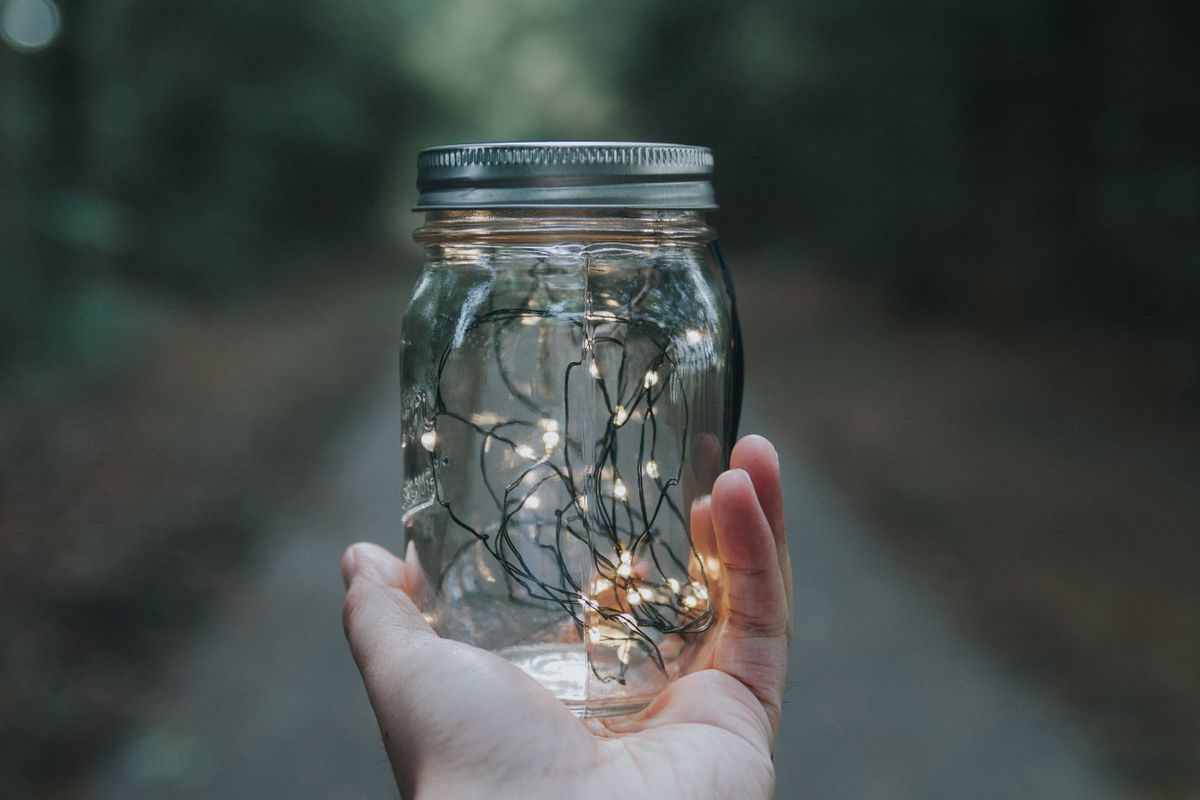 A hand holds a jar filled with glowing fairy lights on a blurred outdoor path.