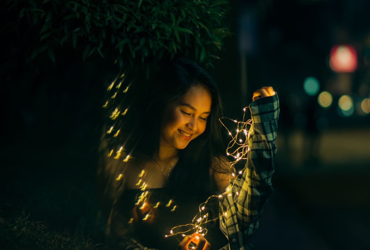 Smiling young woman holding fairy lights at night, creating a warm and magical atmosphere.