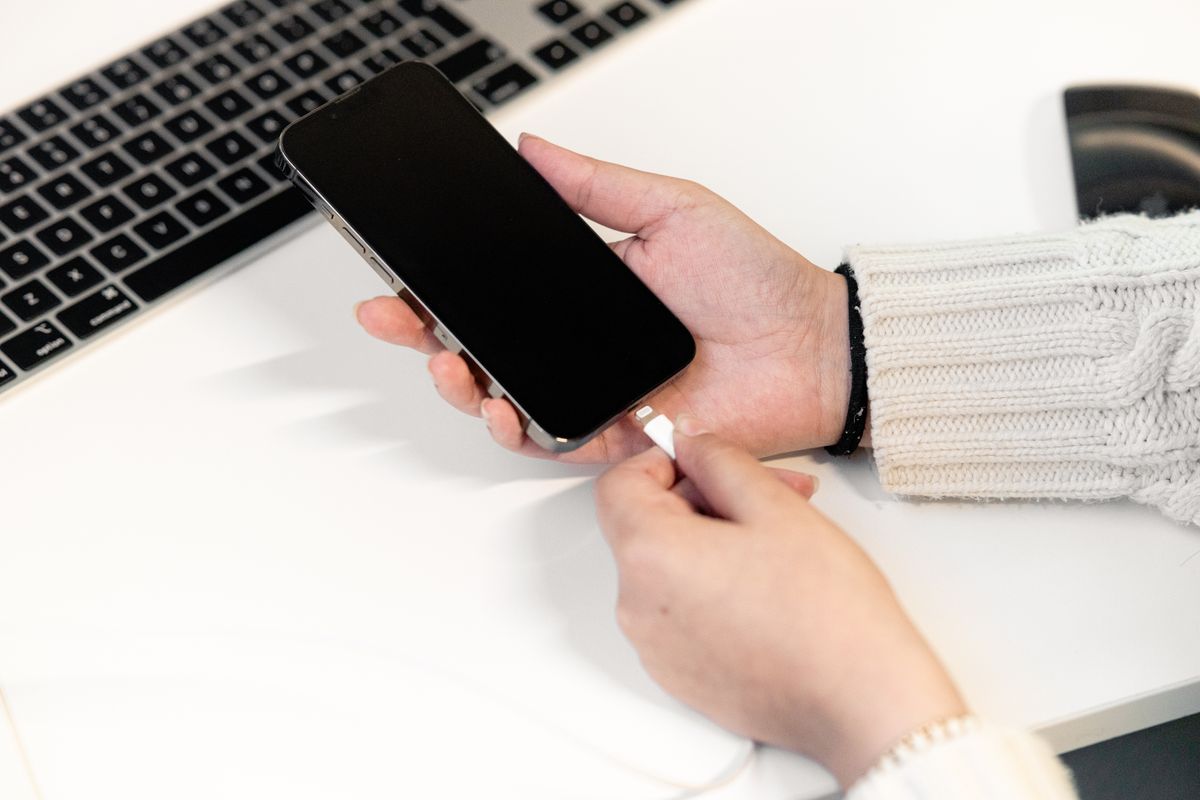 A woman plugs her smartphone into a charger at a desk, next to a laptop, illustrating modern technology usage.