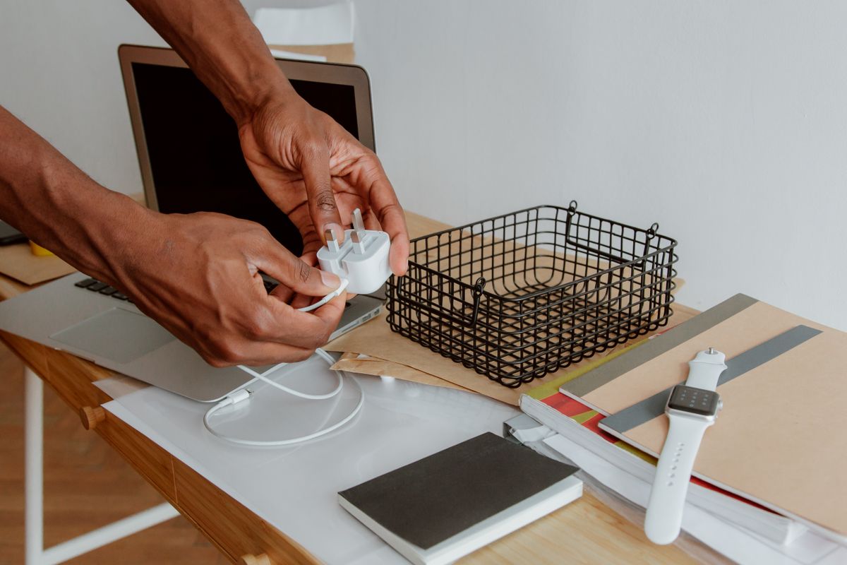 Close-up of a man organizing a desk, handling a charger next to a laptop, wire basket, and notebooks.