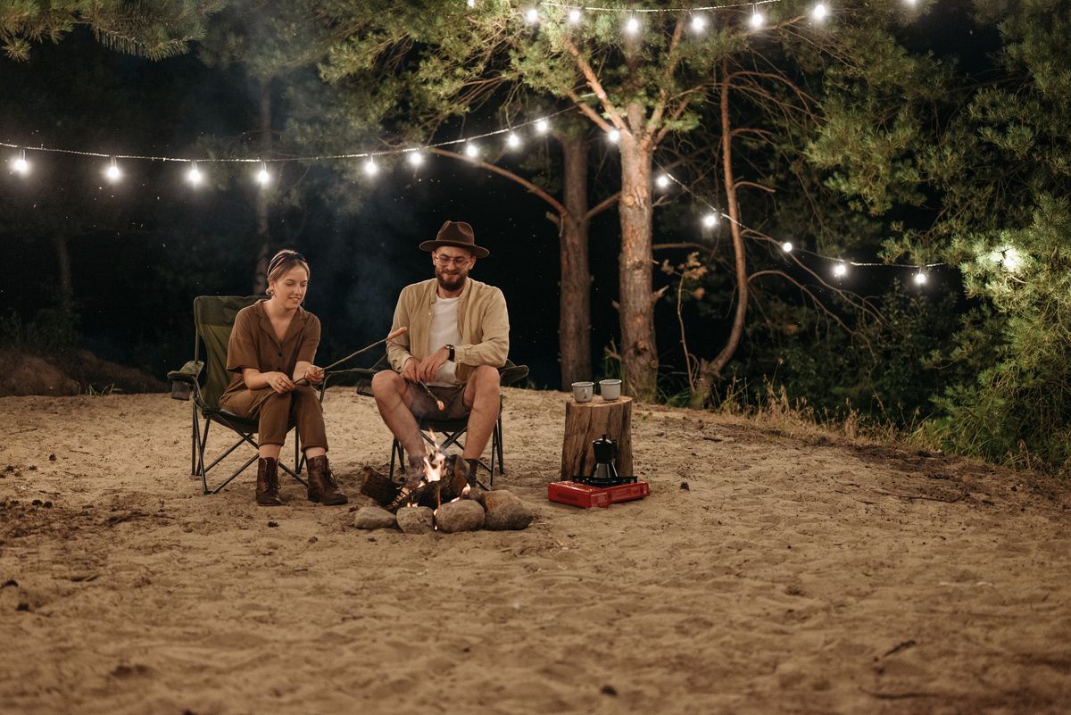 A couple enjoys a peaceful night by the campfire in a forest, surrounded by string lights.
