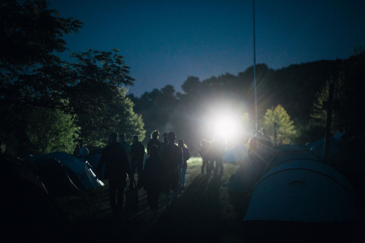 A group of people enjoying a serene camping night illuminated by distant lighting, surrounded by tents and nature.