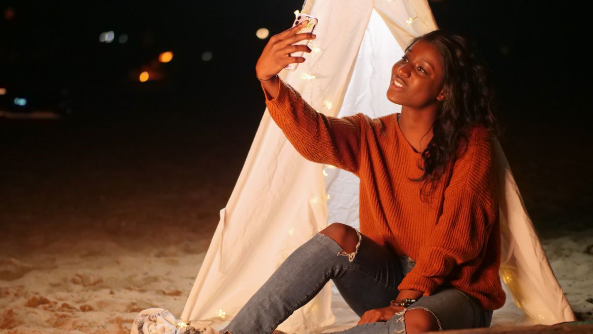 A woman enjoys a nighttime beach camp, taking a selfie by a tent adorned with string lights.