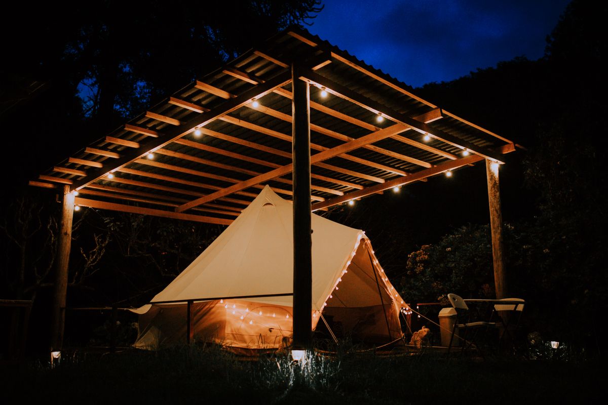 A warmly lit camping tent under a wooden shelter during nightfall.