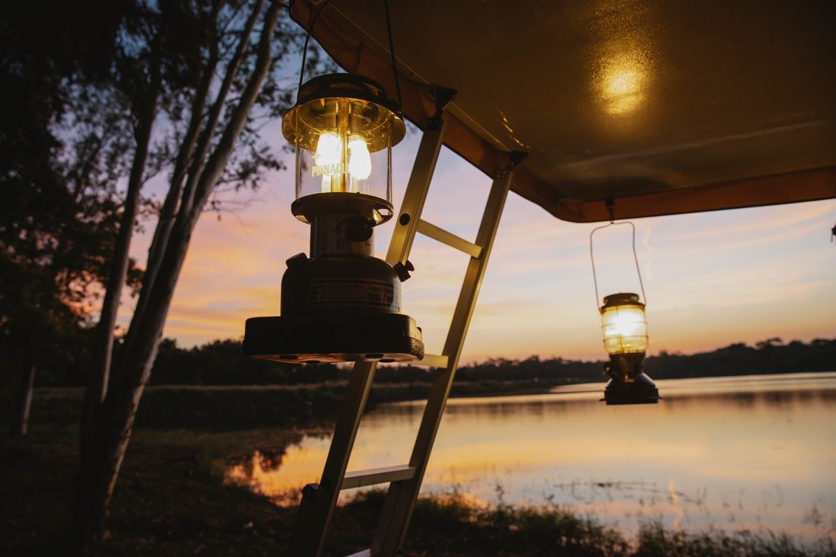 Empty space under camping tent with lamps and stairways placed on coast of lake with thin trees in evening on sunset
