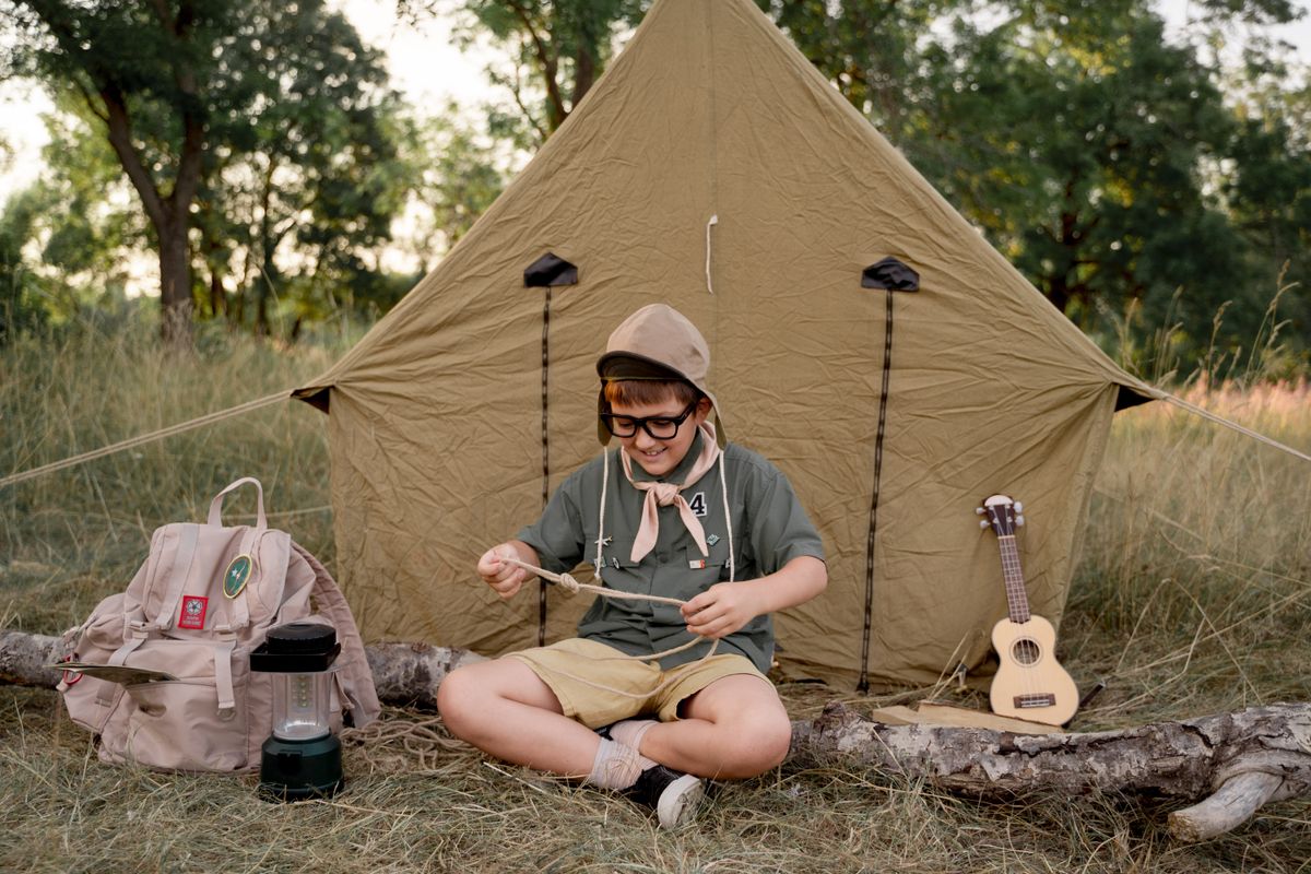 A cheerful young boy in scout uniform ties knots outside his tent on a sunny camping day.
