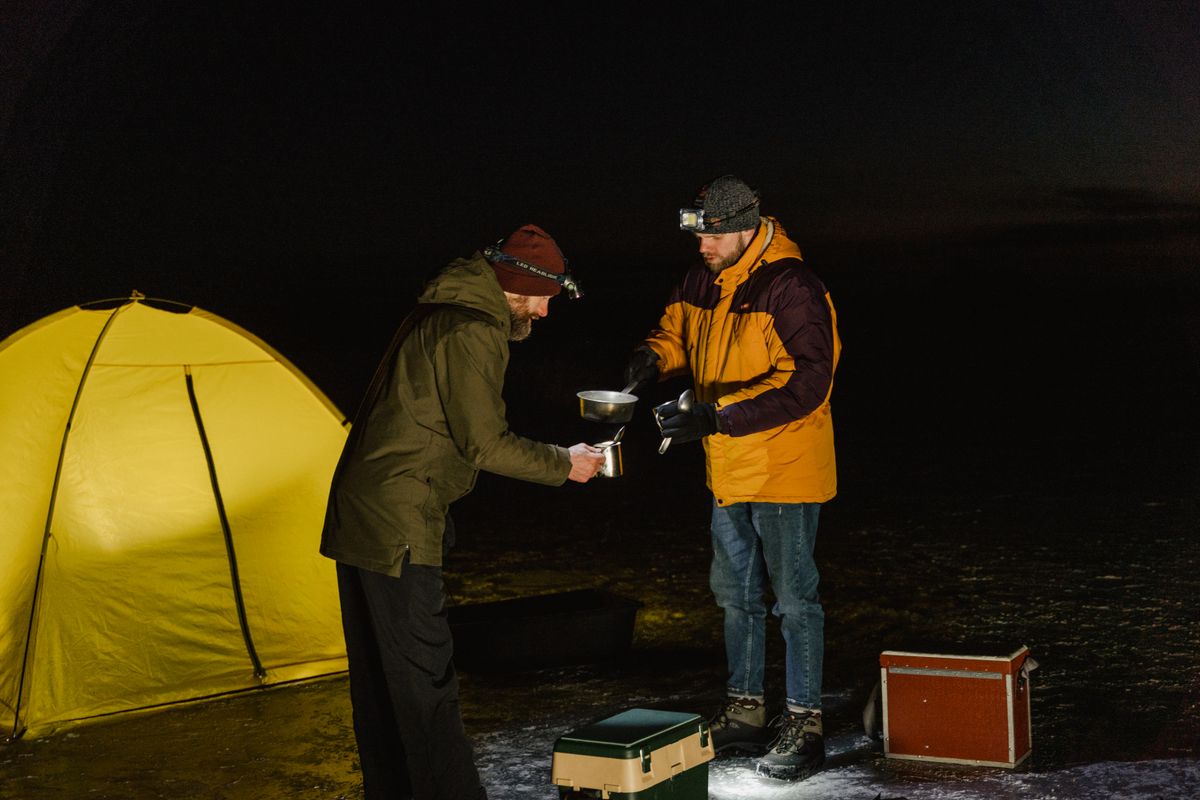 Two men camping at night in winter, sharing a meal by their tent.