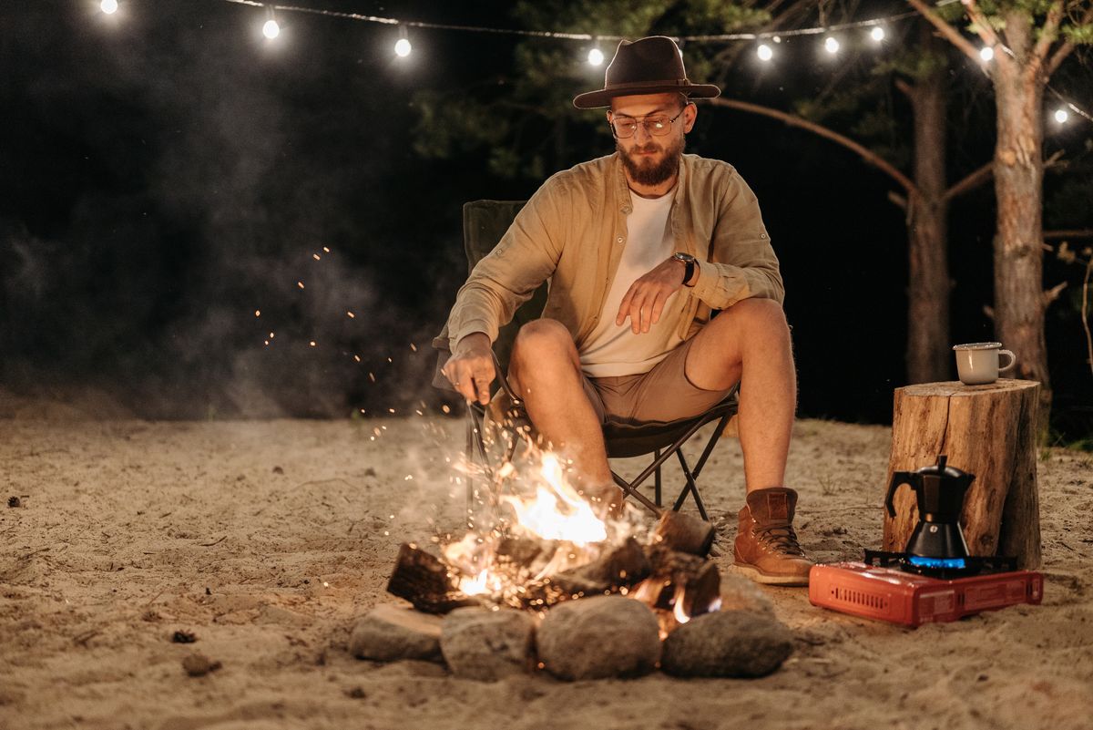 A bearded man in a fedora relaxes by a campfire under string lights at night.