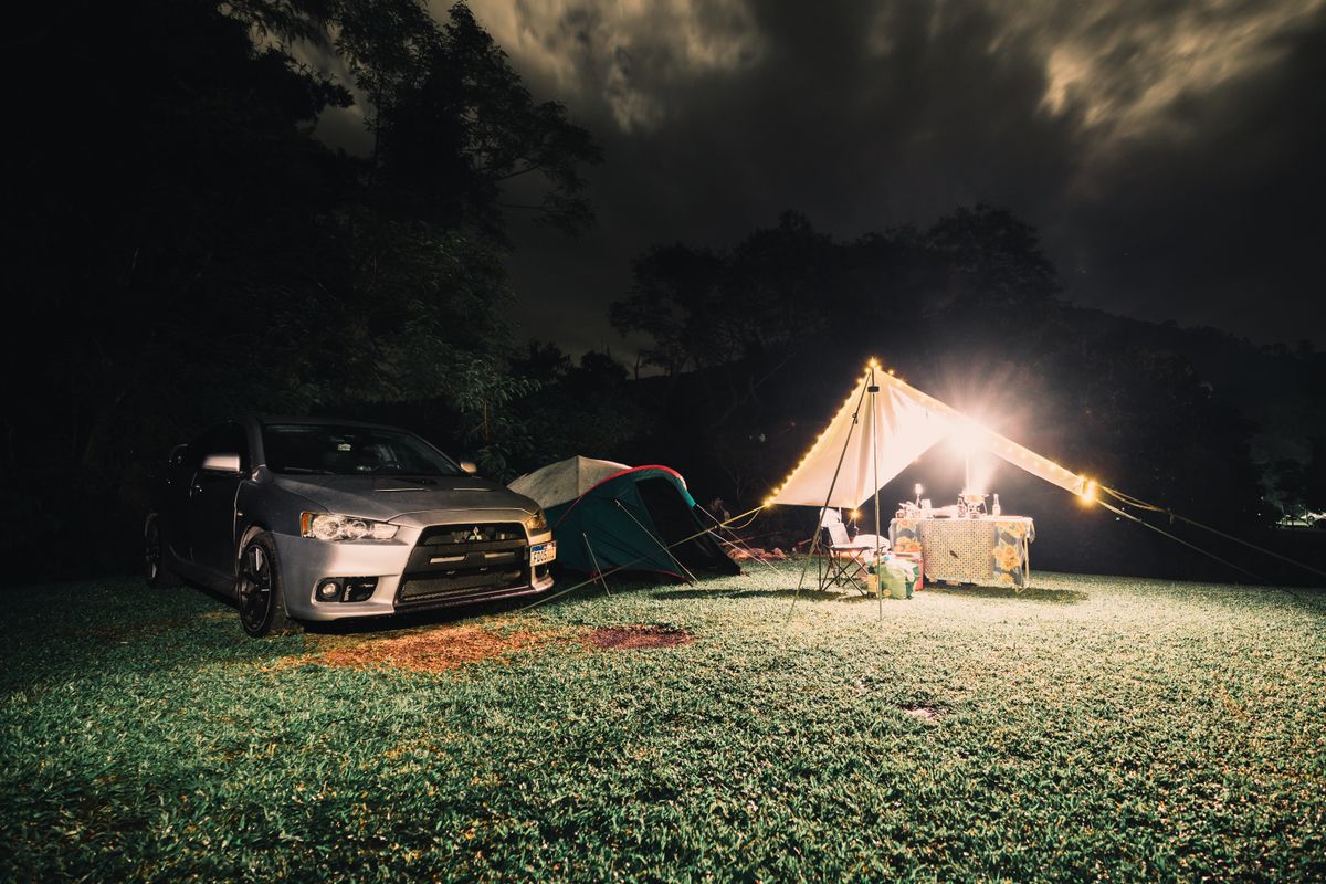Serene camping site at night featuring illuminated tents, car, and lush grass under a cloudy sky.