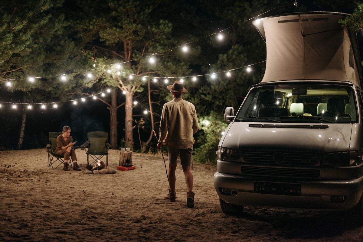 Couple enjoying a camping trip at night with a campervan and string lights illuminating the site.