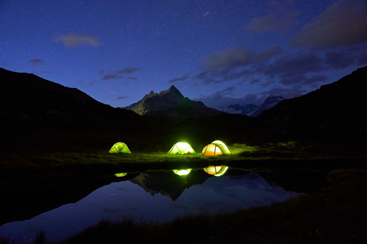 A tranquil night camping by a mountain lake with glowing tents under a starlit sky.