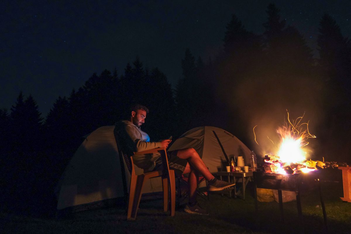 A man enjoys a night camping by the fire, using his smartphone beside a tent under the stars.