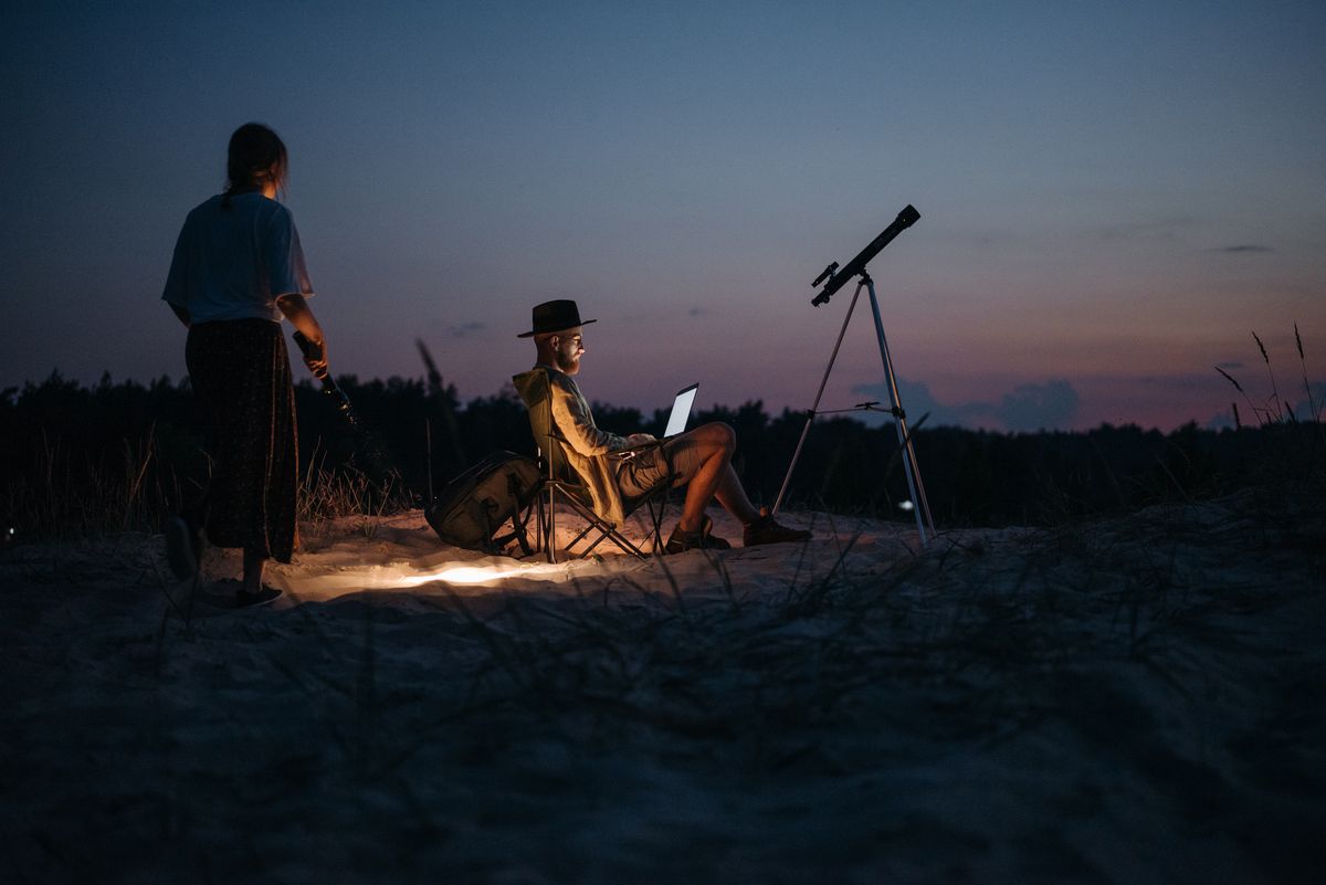 Couple enjoying the night sky at a campsite with telescope and laptop, capturing serene outdoor vibes.