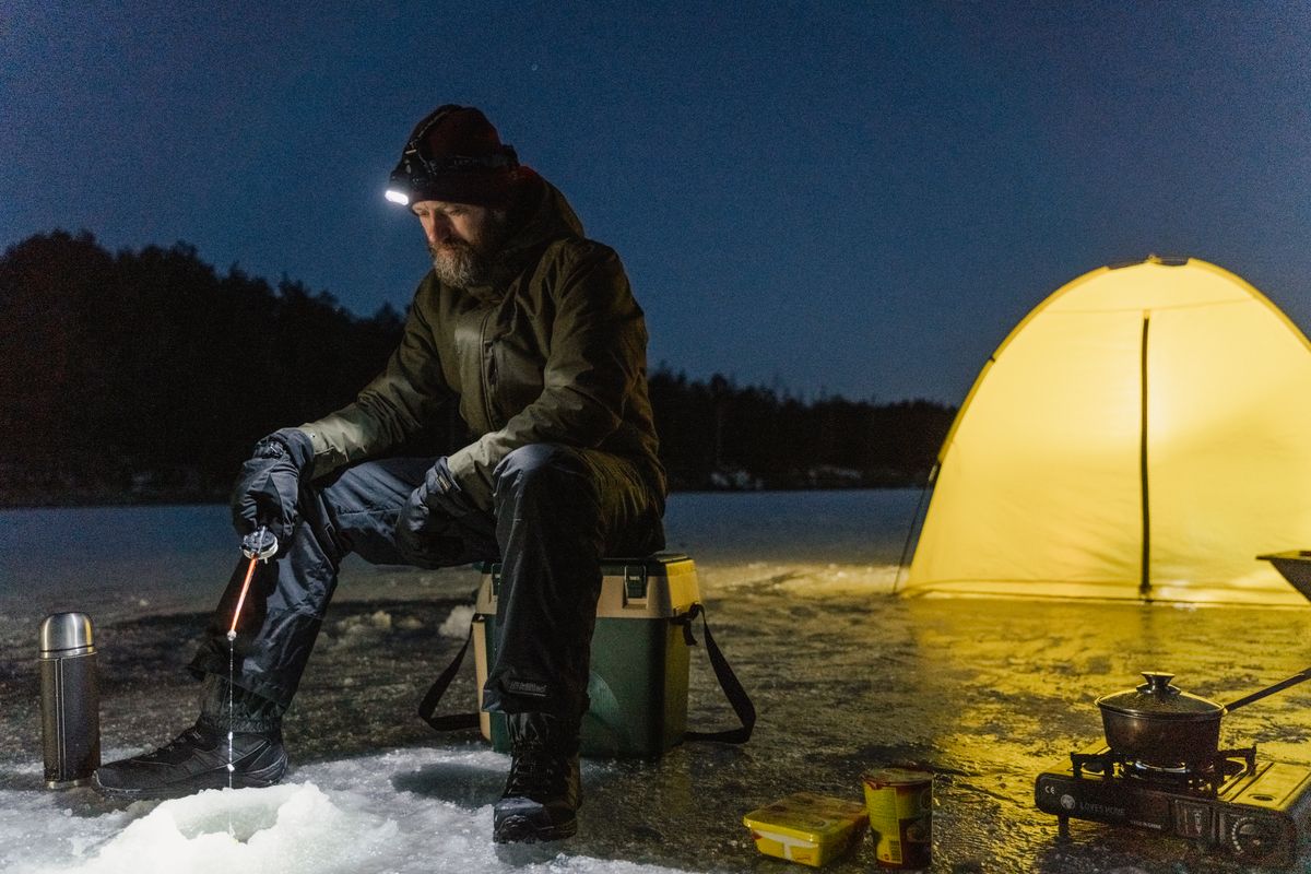 A man ice fishing at night next to a glowing tent on a frozen lake.
