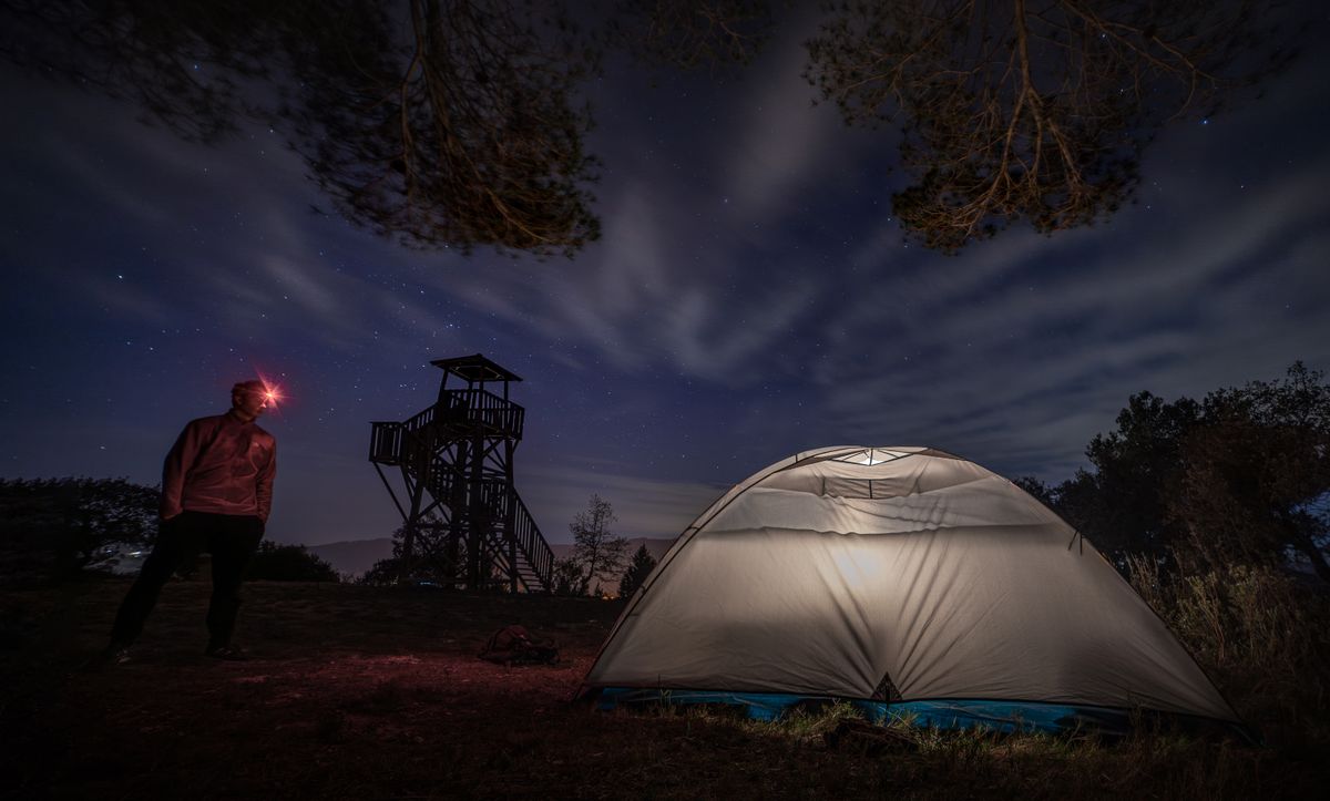 Night scene of a lit tent and person near a watchtower under a starry sky.
