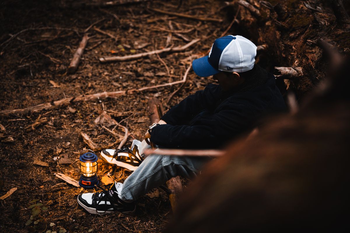 A man wearing a cap sits by a lantern in a forest, creating a serene outdoor atmosphere.