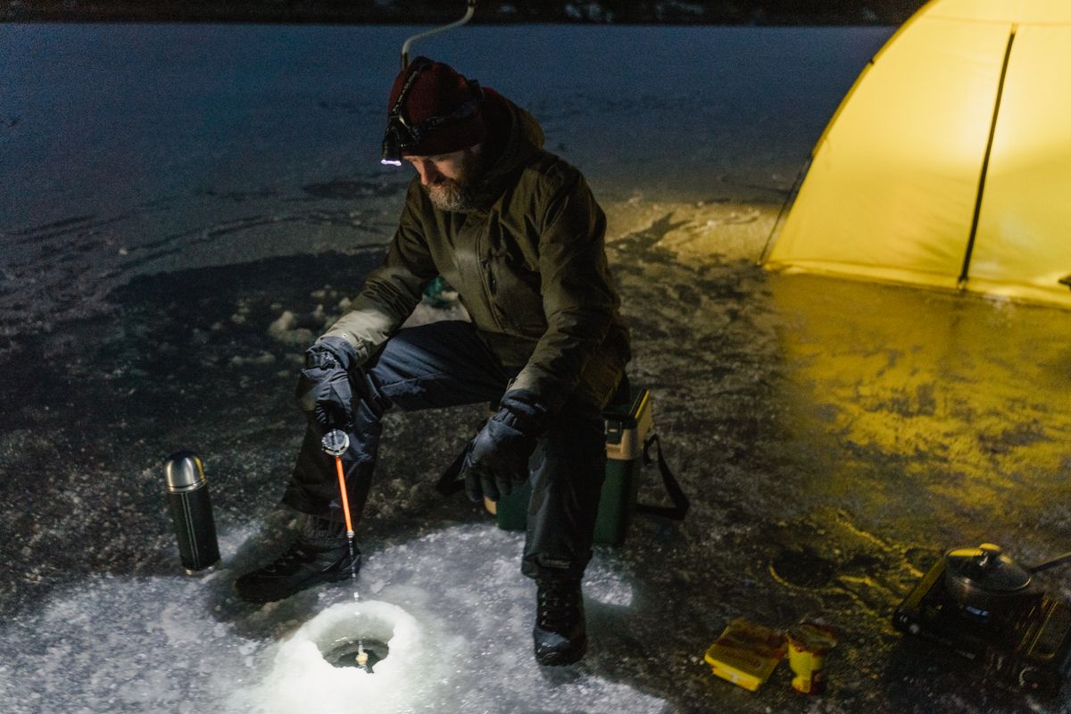 A lone fisherman enjoys ice fishing on a frozen lake at night, illuminated by a camping light.
