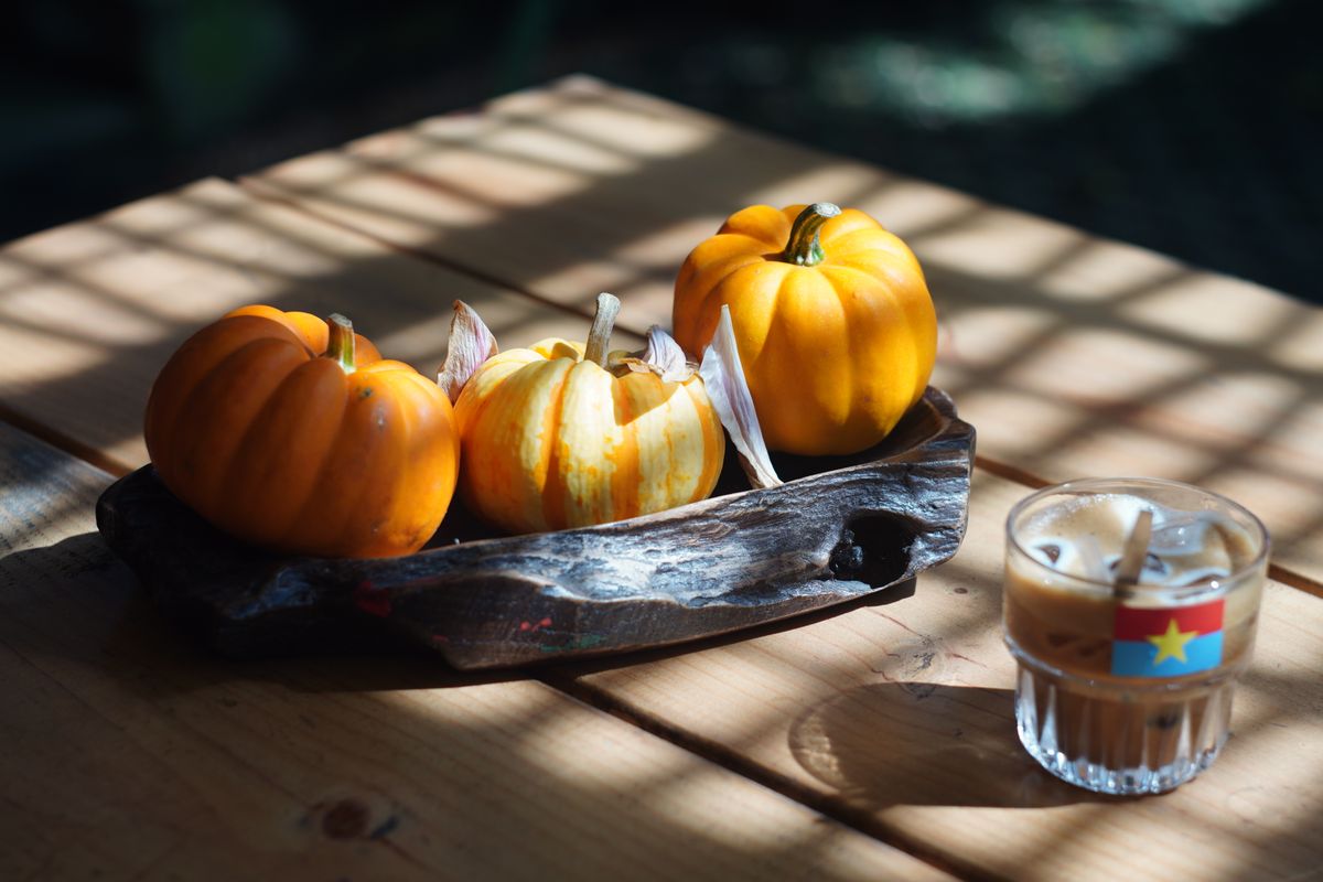 A warm autumn setting featuring mini pumpkins and a glass of coffee on a sunlit wooden table.