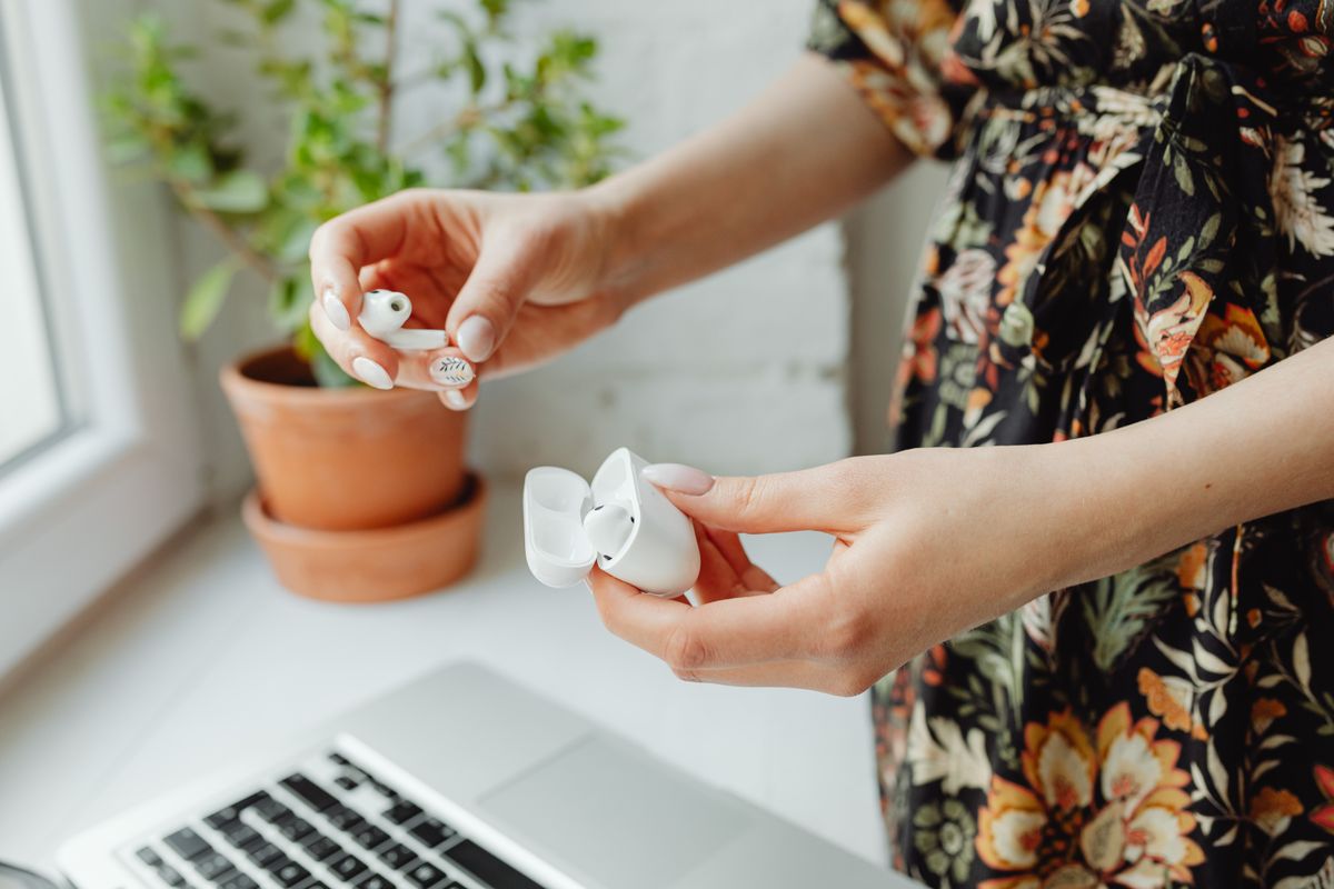 A person holding wireless earbuds and a charging case near a laptop, with a plant in the background.