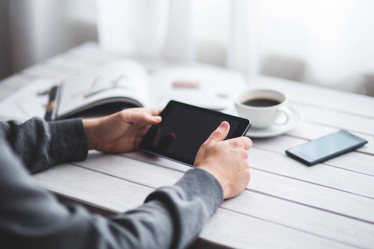 Adult holding a tablet at a minimalist workspace with coffee and a smartphone nearby.