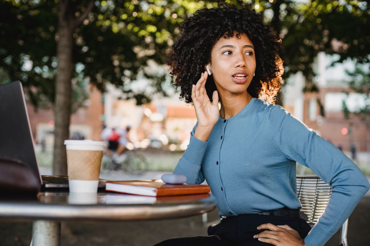 Young woman with afro hair using wireless earbuds at an outdoor cafe, table with tech and coffee.