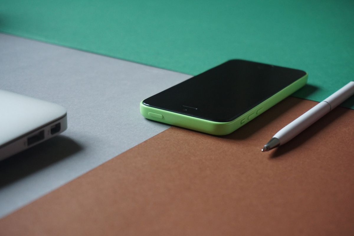 A minimalist workspace featuring a green smartphone, white laptop, and pen on a colored surface.
