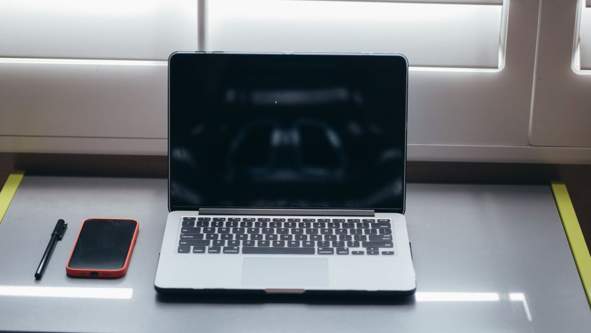 A minimalist desk setup featuring a laptop, smartphone, and pen under soft window light.