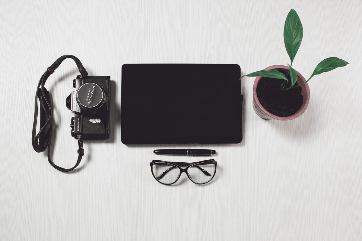 An overhead view of a neat desk setup featuring a camera, tablet, glasses, pen, and plant, emphasizing modern work aesthetics.