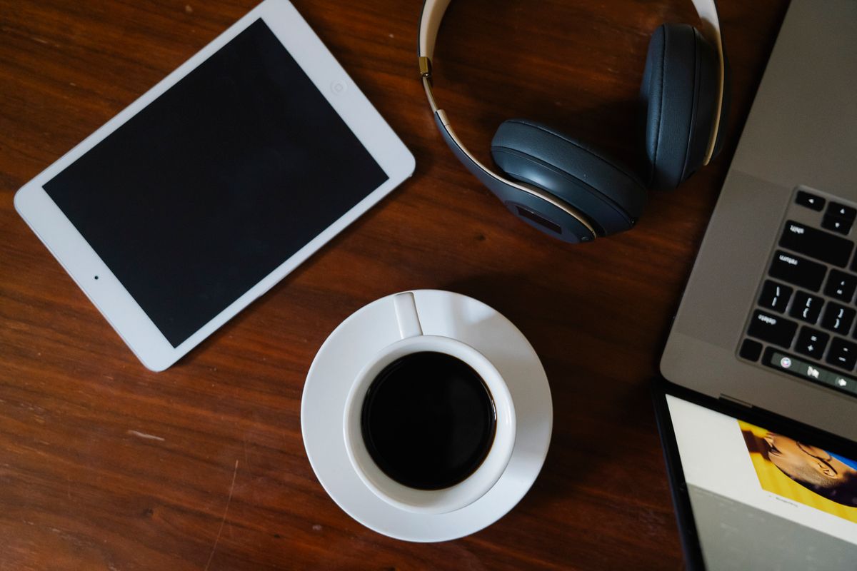 Overhead shot of a coffee cup, laptop, tablet, and headphones on a wooden desk.